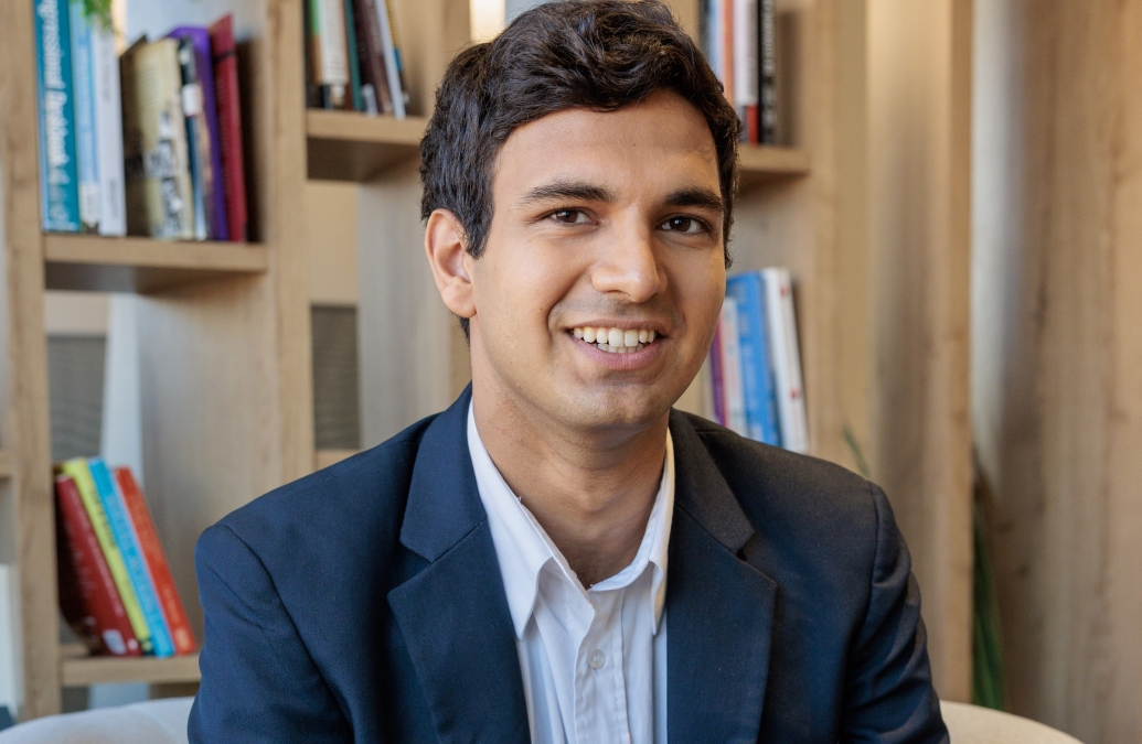 head and shoulder portrait of a smiling young man in a suit in front of books.