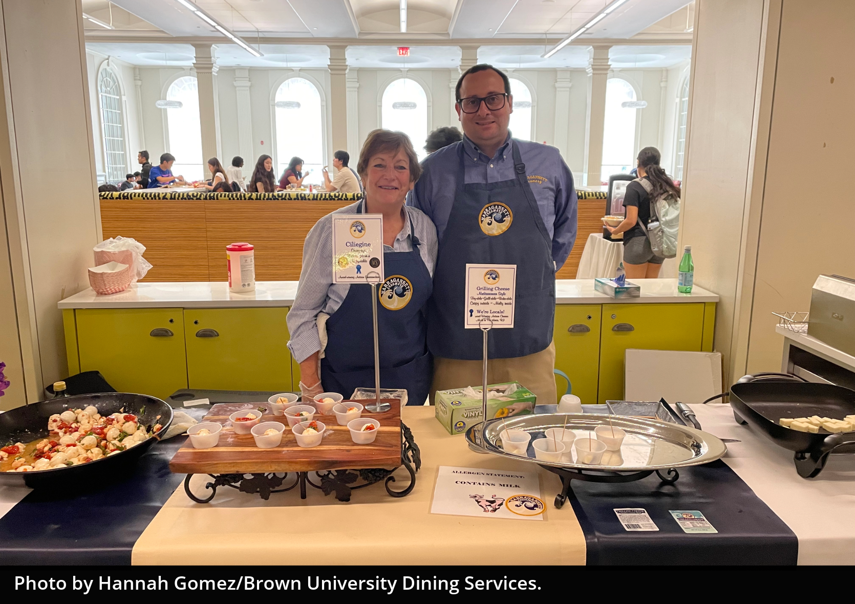 Two people wearing Narragansett Creamery aprons smile at the camera behind a table holding a tray of cheese they are serving.