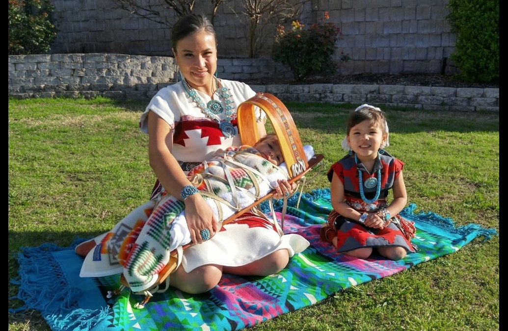 three native americans on a colorful blanket outdoors on green grass in the sun.  One adult, one child and one baby dressed in customary garb.