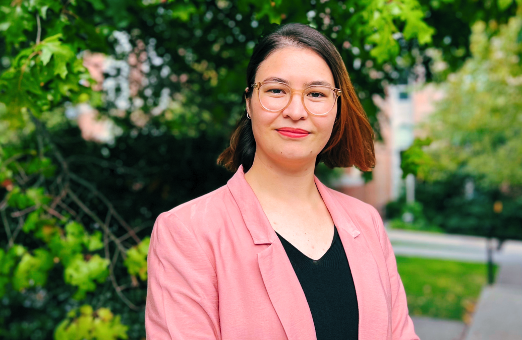 head and shoulders portrait of a young person wearing glasses and a pink blazer in an outdoor setting.