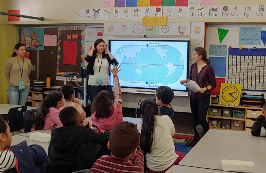 A student raising her hand in a classroom with three teachers at the front