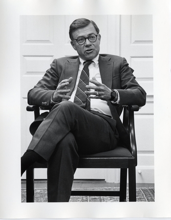 Black and white photo of seated figure of howard swearer in a suit and tie, round glasses and gesturing as he speaks. 