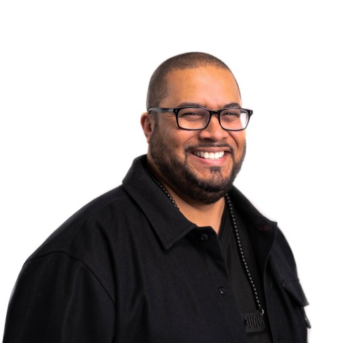 Headshot of Juan Wilson Jr. smiling in a black shirt standing to the side in front of a white background.