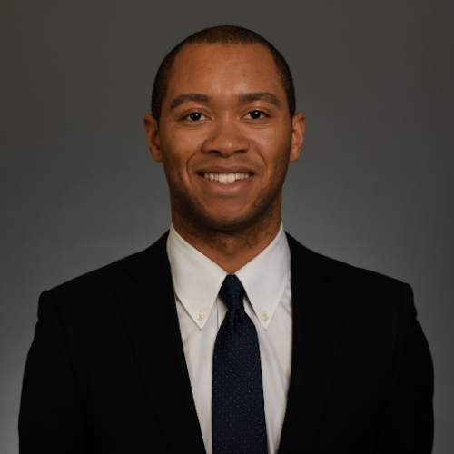 Headshot of Lloyd Ocean smiling at the camera in a black suit against a neutral gray background.