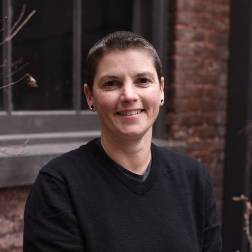 Headshot of Michelle Duso wearing a black sweater, standing in front of a building exterior and smiling.