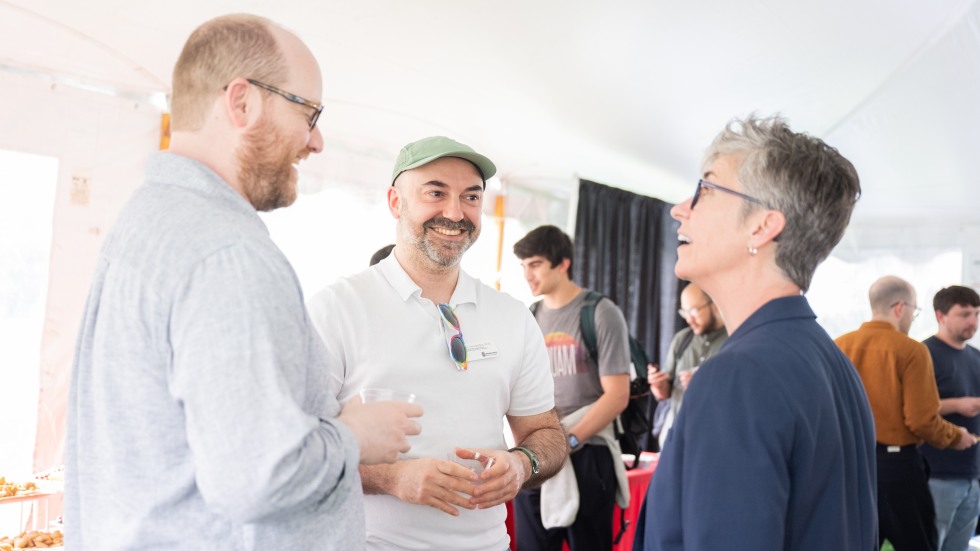 Candid photo of VP Mary Jo Callan, Director jesús hernández and community member talking, standing and smiling underneath a tent with other people socializing in the background. 