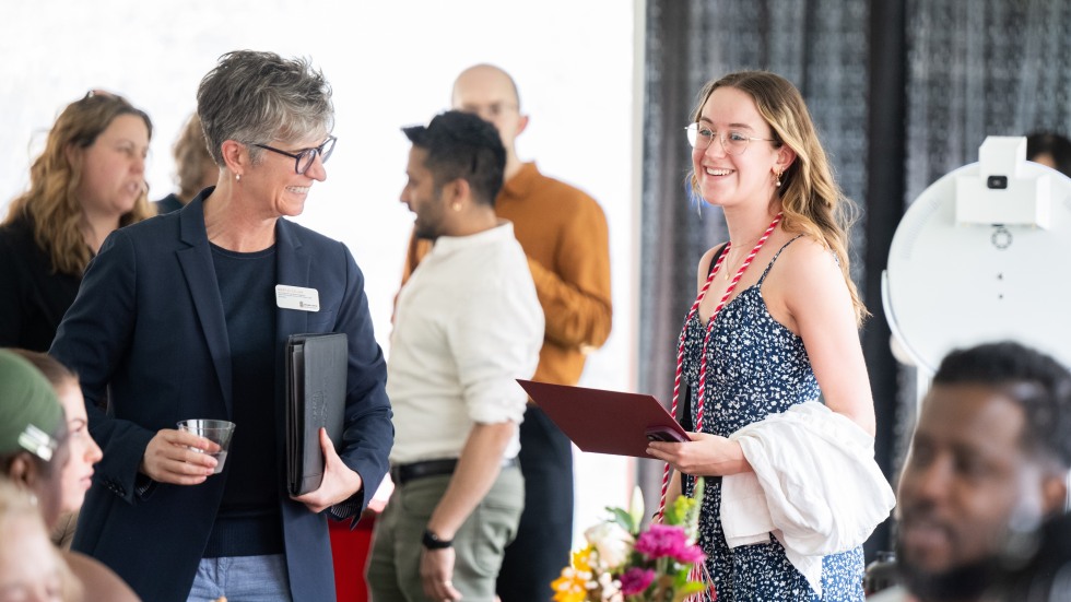 Candid photo of VP Mary Jo Callan and student award winner Elizabeth (Ellie) Morvatz talking and smiling, standing near others seated at a table. 
