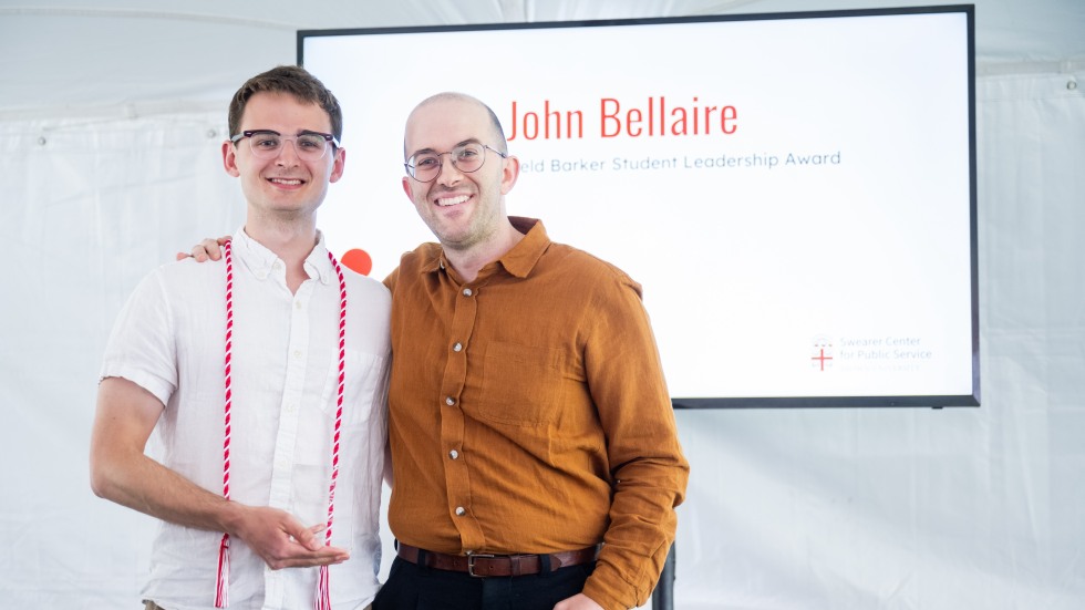 Photo of student award winner John Bellaire smiling and posing with presenter Austin Wilson, a digital slide depicting his name and the award name is their backdrop.
