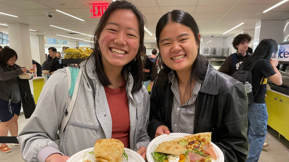 Two students smiling at the camera, holding up plated sandwiches. 