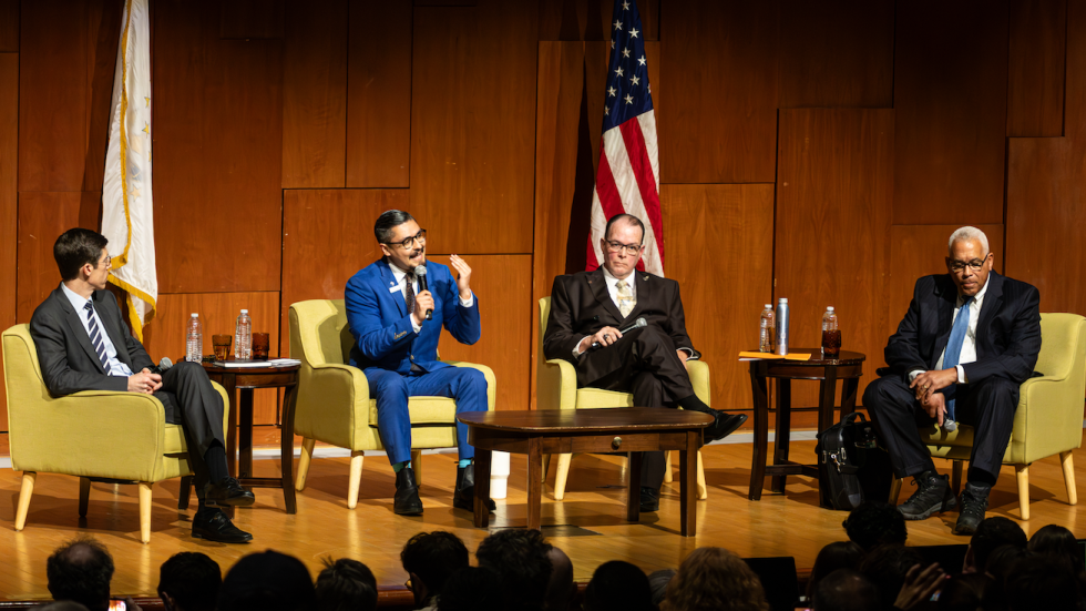 Four Providence mayoral candidates sit on stage during a public forum at Brown University, seated side by side in chairs with microphones as they participate in a moderated discussion.