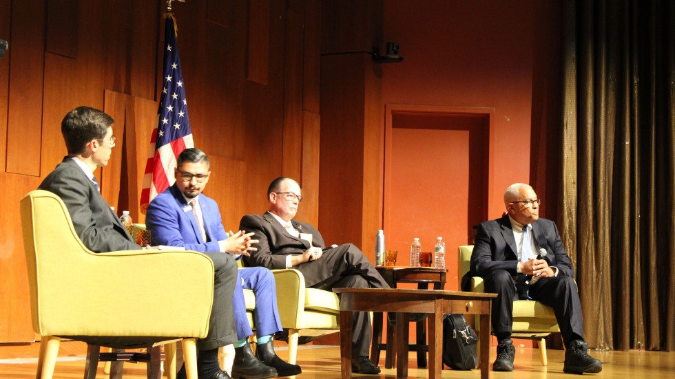 Four Providence mayoral candidates sit on stage during a public forum at Brown University, seated side by side in chairs with microphones as they participate in a moderated discussion.